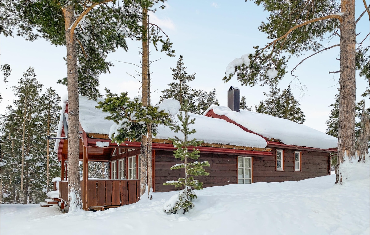 A charming wooden vacation home is surrounded by a snowy landscape, with trees providing a natural backdrop. A thick layer of snow covers the roof and ground, creating a serene winter atmosphere. The structure features a welcoming porch, inviting guests to explore the outdoors.