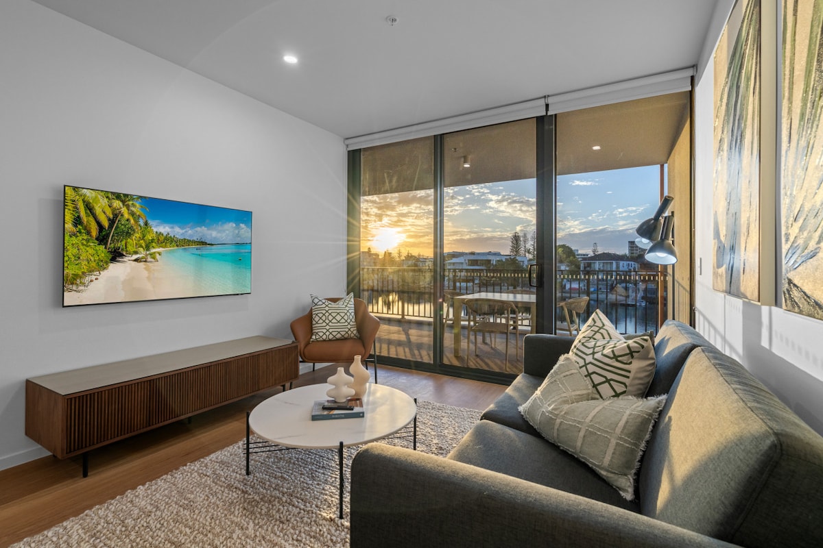 A spacious living area features a comfortable sofa and a modern coffee table. A large smart TV is mounted on the wall. Floor-to-ceiling glass doors offer views of the sunset and waterfront, allowing natural light to fill the room.