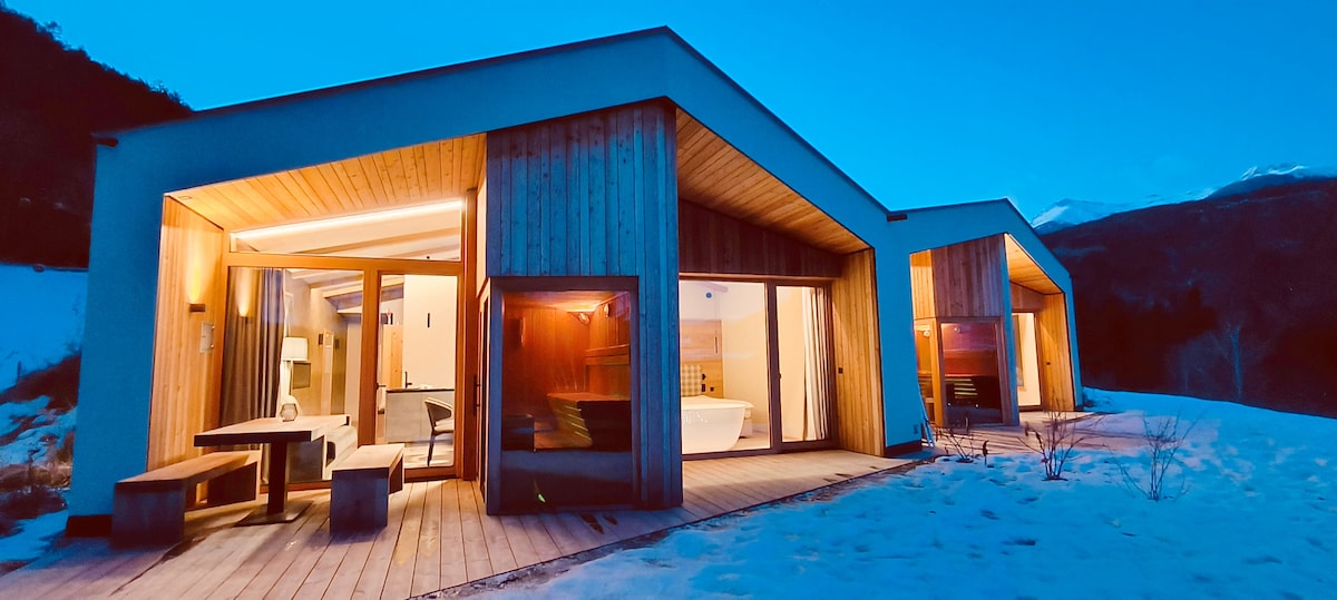 The exterior of the chalet features a modern design with wooden paneling and large glass doors, allowing for ample natural light. A snow-covered landscape surrounds the building, while soft lighting highlights the structure during dusk.