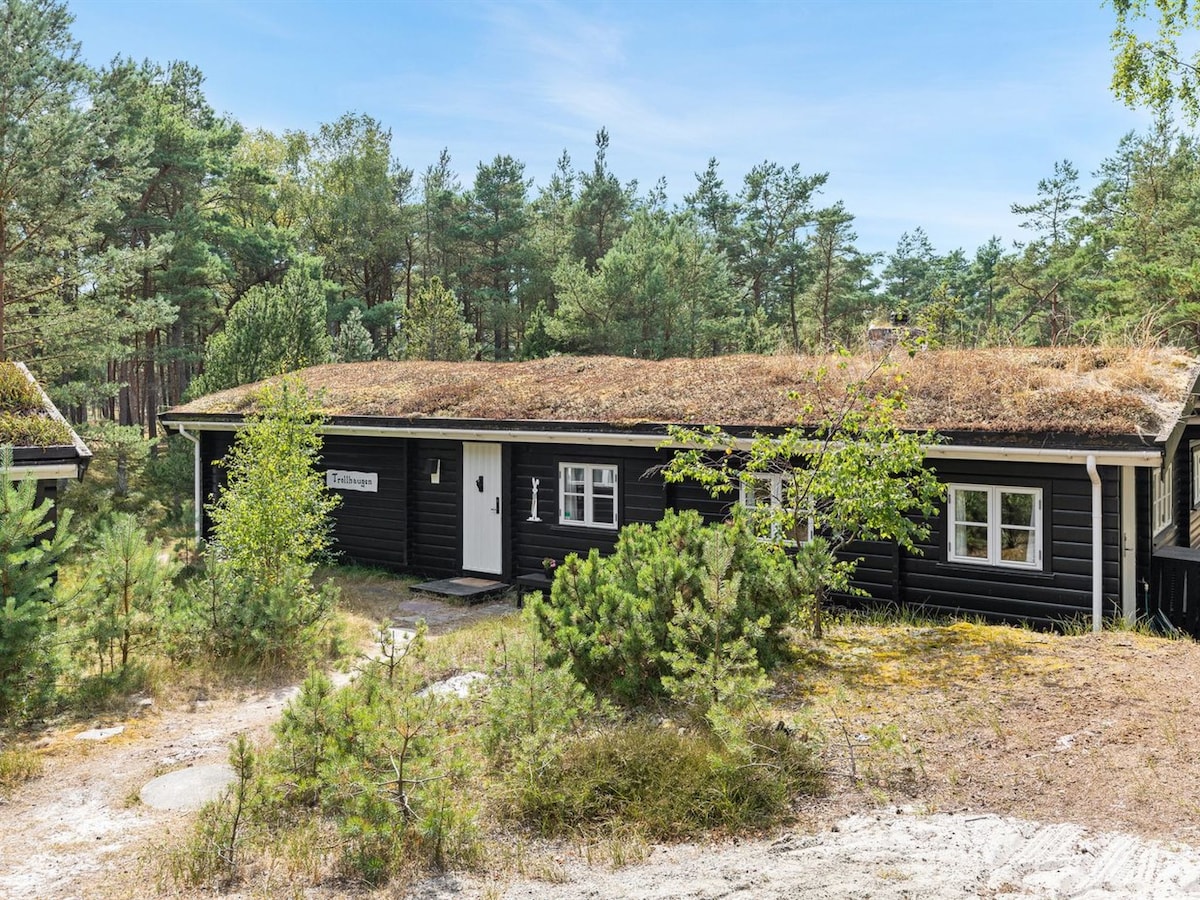 A single-family house with a natural grass roof is surrounded by trees. The structure features white window frames and a welcoming front door. Pathways lead through the natural landscape, emphasizing a blend of rustic architecture and the surrounding greenery.