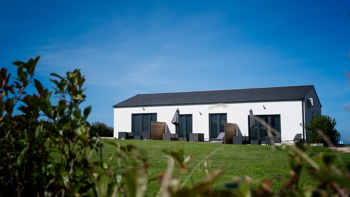 A modern single-story building is shown with a sleek design. Outdoor seating areas are visible, featuring umbrellas for shade. The lush green lawn surrounds the property, enhancing the tranquil setting under a bright blue sky.