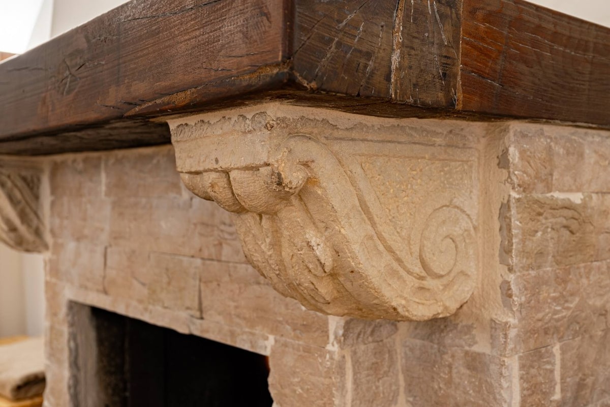A detailed view of a rustic stone fireplace is highlighted, showcasing an intricately carved corbel supporting a wooden mantle. The natural stone texture and soft finish contribute to the warmth of the space, inviting relaxation and comfort.