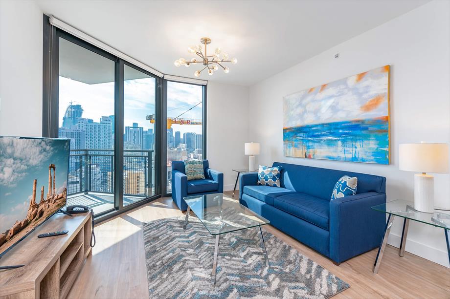 A contemporary living area features floor-to-ceiling windows offering sweeping city views. A blue sofa and armchair are arranged around a glass coffee table, while modern lighting fixtures illuminate the space. A television is positioned on a wooden media console.