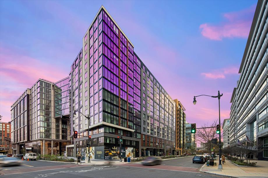 A modern building complex displays an impressive facade of glass and concrete, reflecting hues of purple and pink from the sunset sky. The street-level features large windows and storefronts, while the surrounding area includes traffic lights and urban landscaping.