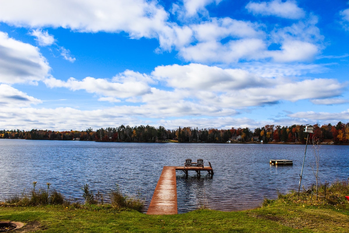 A private dock extends over the tranquil waters of Chain Lake, framed by a colorful landscape of trees. Two chairs are positioned on the dock, overlooking the lake. A distant platform adds to the serene view under a partly cloudy sky.