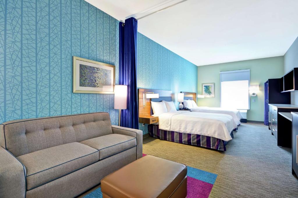 A spacious hotel room features two queen beds with soft bedding against a patterned blue wall. A gray sofa and an ottoman are situated in front of the beds, providing additional seating. Natural light fills the room from a window adorned with light-filtering shades.
