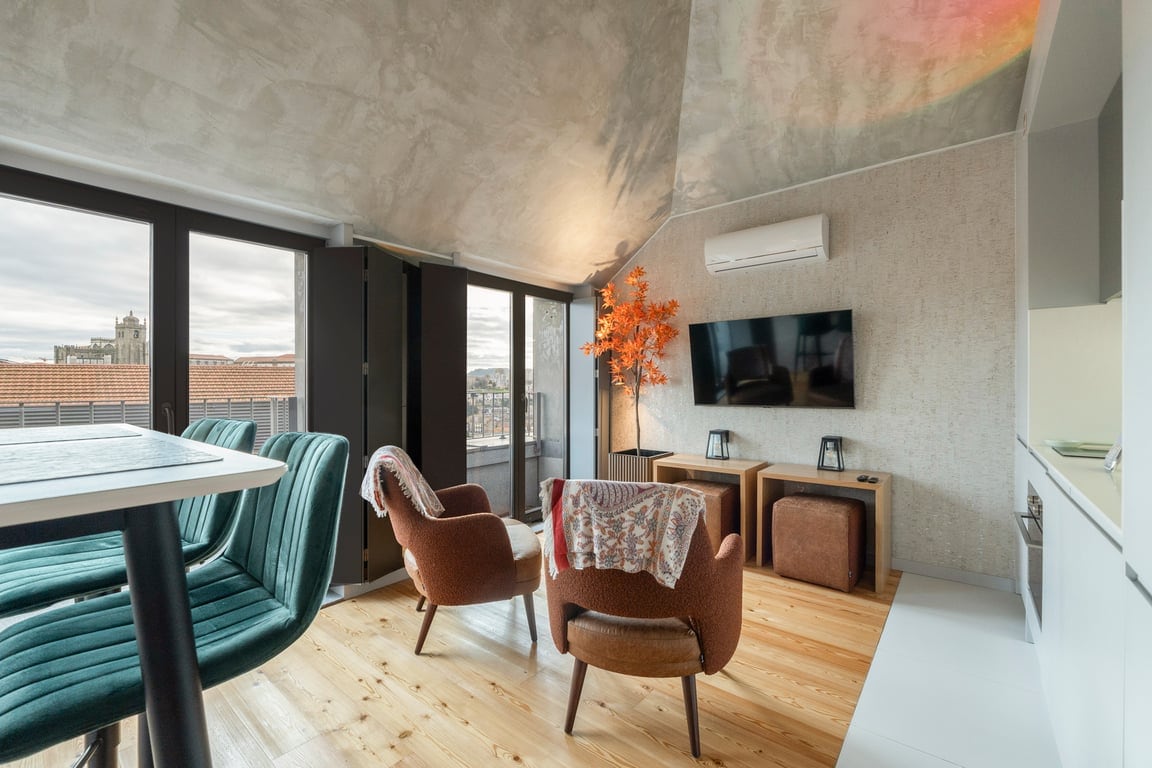 A modern living area features a high table with tall chairs and two comfortable lounge chairs positioned nearby. A flat-screen TV is mounted on the wall, complemented by decorative wooden furniture. Large windows provide natural light and showcase views of the city.
