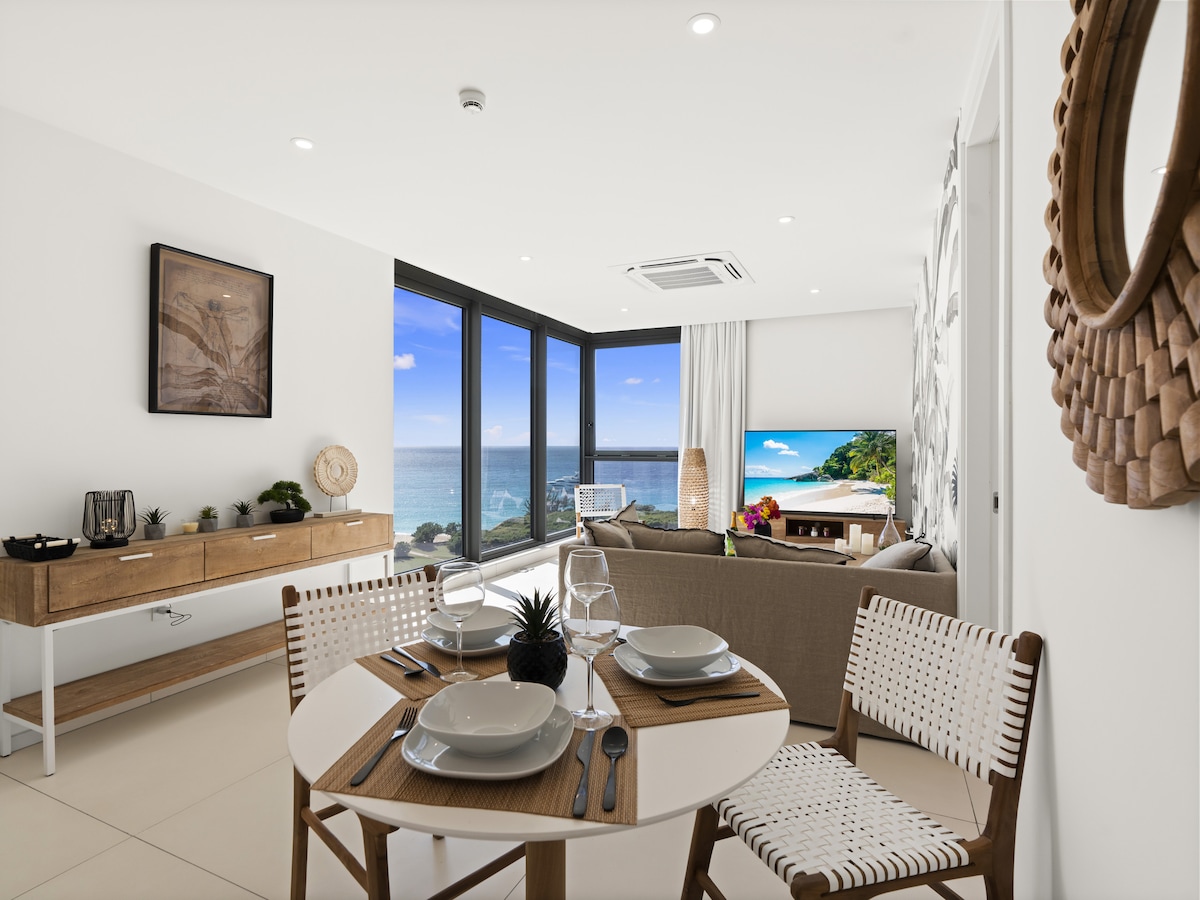 An open-concept living area features a dining table set for four and modern decor. Large windows provide a view of the beach and ocean, while natural light brightens the space. A comfortable seating area with a TV enhances the coastal ambiance.