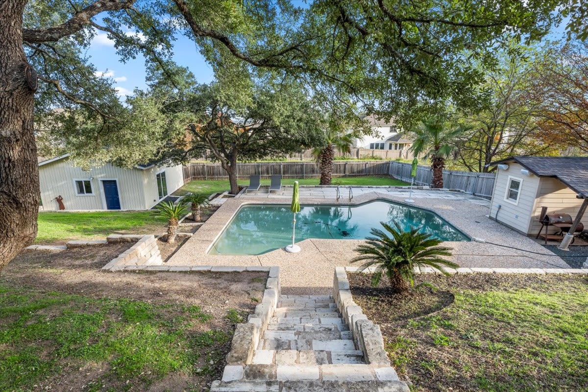 A private pool is set within a spacious outdoor area, surrounded by landscaped greenery and towering trees. Lounge chairs are positioned on the pool deck, offering relaxation options. A steps pathway leads down to the pool area, enhancing accessibility and inviting enjoyment of the space.