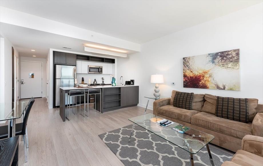 A contemporary living space features a comfortable sofa and glass coffee table, with natural light pouring in from large windows. The kitchen area showcases stainless steel appliances, bar seating, and modern cabinetry. A vibrant piece of wall art adds a touch of character to the neutral-toned decor.