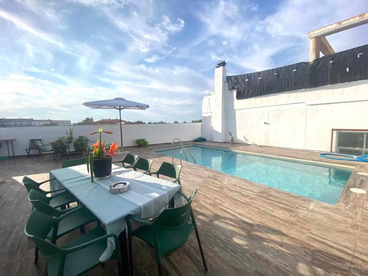 A spacious outdoor area features a rectangular swimming pool surrounded by wooden decking. A large table set for dining accommodates multiple chairs. An umbrella provides shade, while potted plants add greenery to the setting. Clear blue skies complement the relaxing environment.