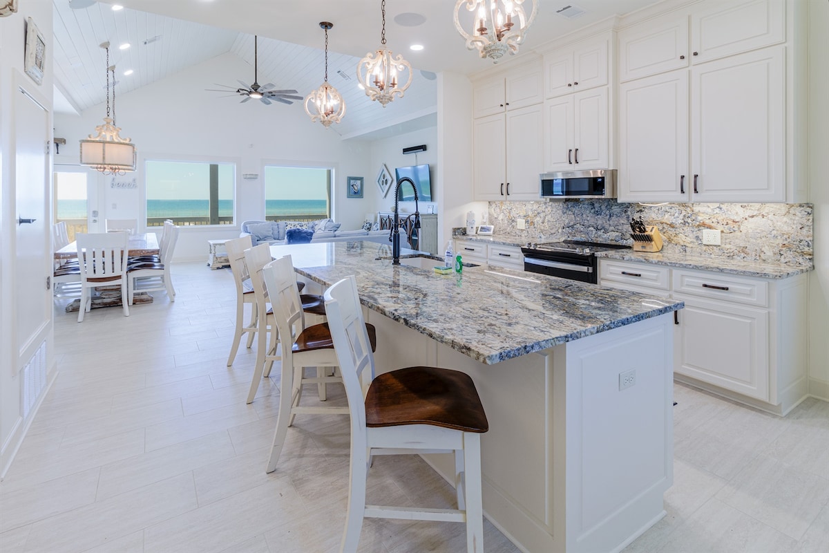 An open kitchen and dining area features light-colored cabinetry and a large granite countertop with seating for several. Pendant lights illuminate the space. In the background, coastal decor can be seen in the living room, paired with expansive windows offering views of the Gulf.