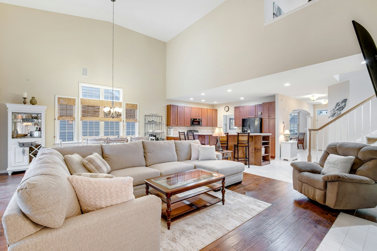 The open living area features a large sectional sofa and a glass-top coffee table, complemented by natural light streaming through multiple windows. A spacious dining area and well-equipped kitchen are visible in the background, enhancing the room's inviting layout.