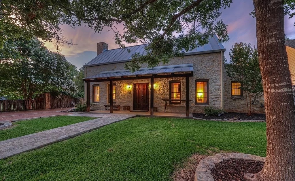 A charming stone house is framed by lush greenery and a well-maintained lawn. Warm light spills from the windows, inviting an atmosphere of comfort. A stone pathway leads to a welcoming front porch adorned with wooden accents beneath a metal roof.