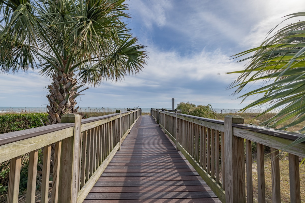 A wooden walkway stretches toward the beach, flanked by lush palm trees. The pathway is lined with railings and intersects with patches of greenery, leading to an open view of the ocean under a clear sky.