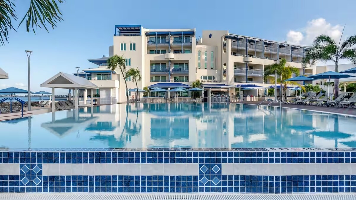 A large swimming pool reflects the clear blue sky and resort building, creating a serene atmosphere. Surrounding the pool are several lounge chairs and umbrellas, providing shaded seating areas. The spacious resort complex features multiple levels and tropical landscaping, emphasizing the welcoming Caribbean setting.