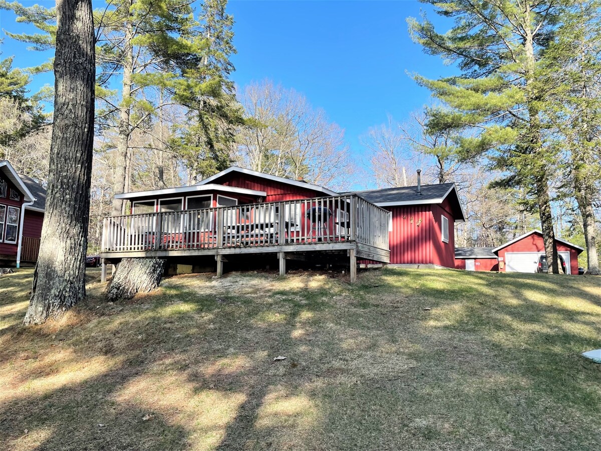 A red cabin is nestled among tall trees on a gentle slope, featuring a spacious deck that extends from the building. A screened-in porch with large windows allows for outdoor enjoyment, while the surrounding landscape showcases a mix of grass and natural vegetation.