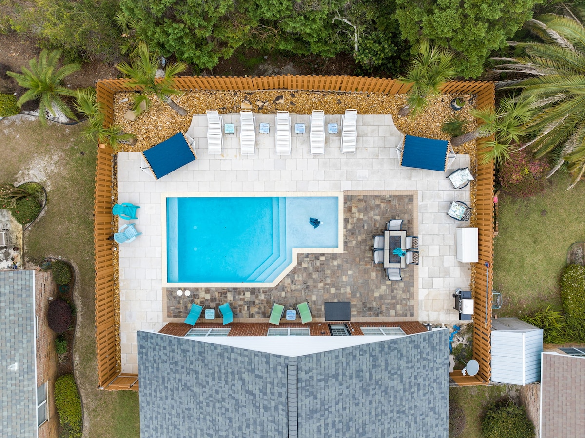 An aerial view of the backyard highlights a spacious saltwater pool surrounded by a tiled area. Several lounge chairs are positioned around the pool, along with seating arrangements on a stone patio. Palm trees and lush greenery frame this serene outdoor space.