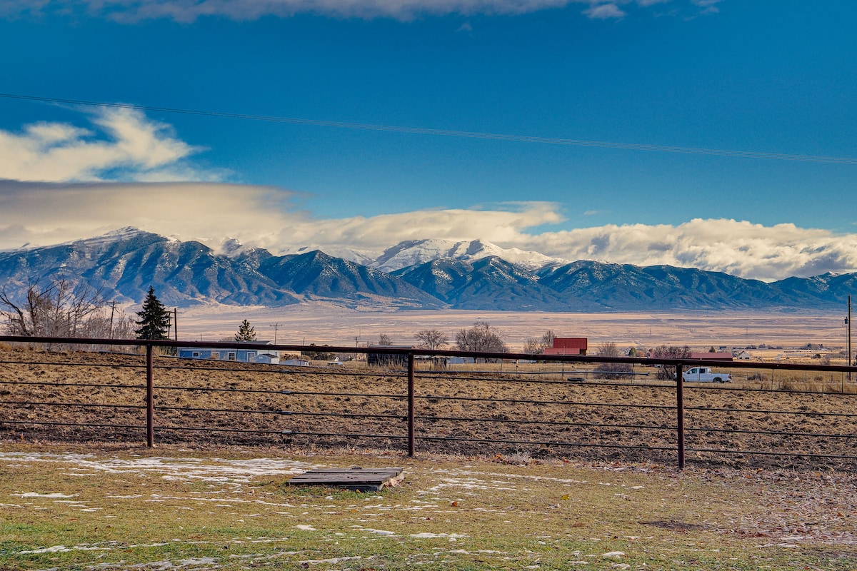 A scenic view captures the expansive Stansbury Mountains under a partly cloudy sky. A rustic fence separates the grassy foreground from the open fields, with hints of nearby structures visible in the distance.