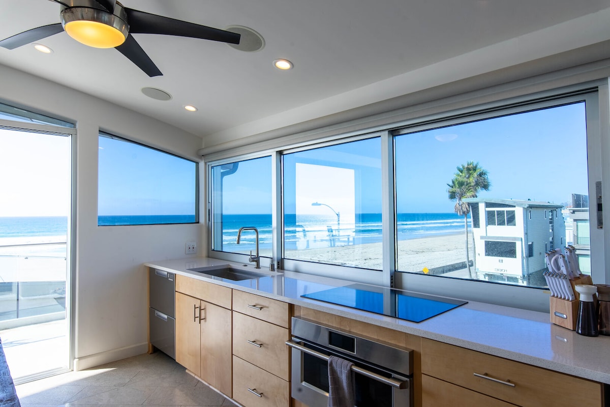 A modern kitchen is showcased with light wood cabinetry and stainless steel appliances. Expansive windows provide unobstructed views of the beach and ocean, allowing natural light to fill the space. A ceiling fan enhances airflow while a sleek countertop offers ample preparation area.