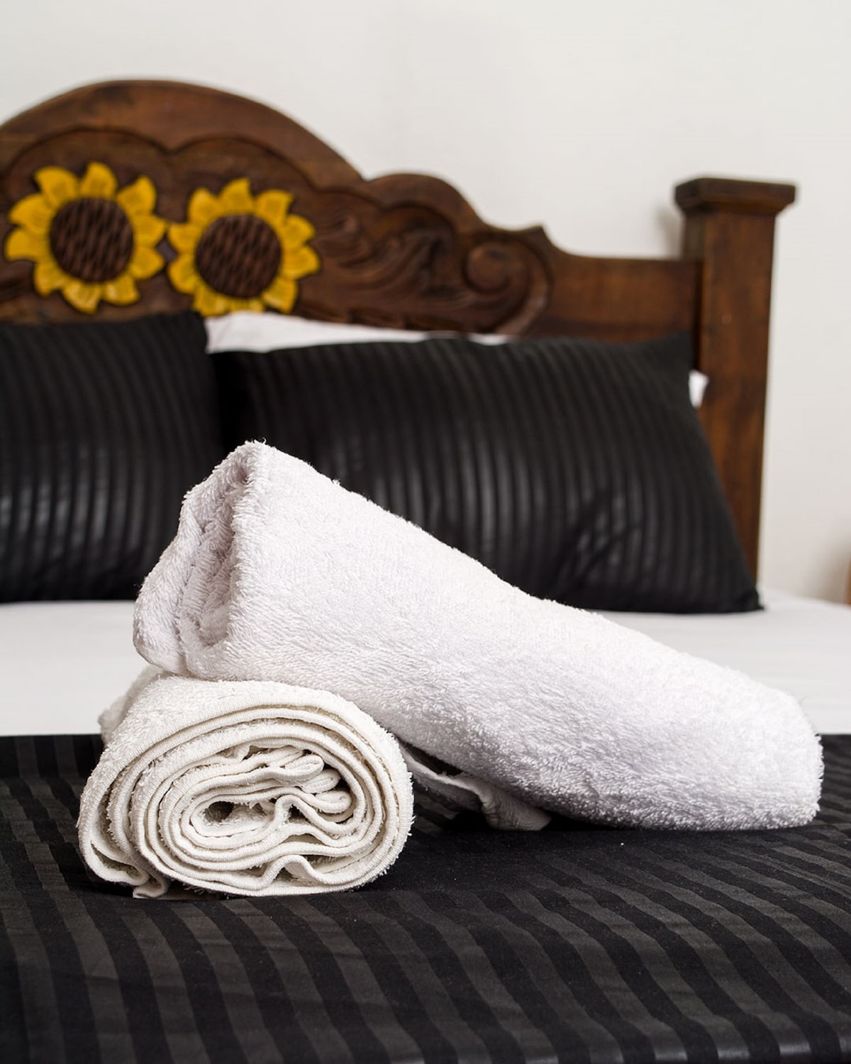 Two neatly arranged towels are presented on a dark striped bedspread. A wooden headboard with sunflower accents frames the spacious bed, which is adorned with textured black pillows, creating a simple yet cozy atmosphere.