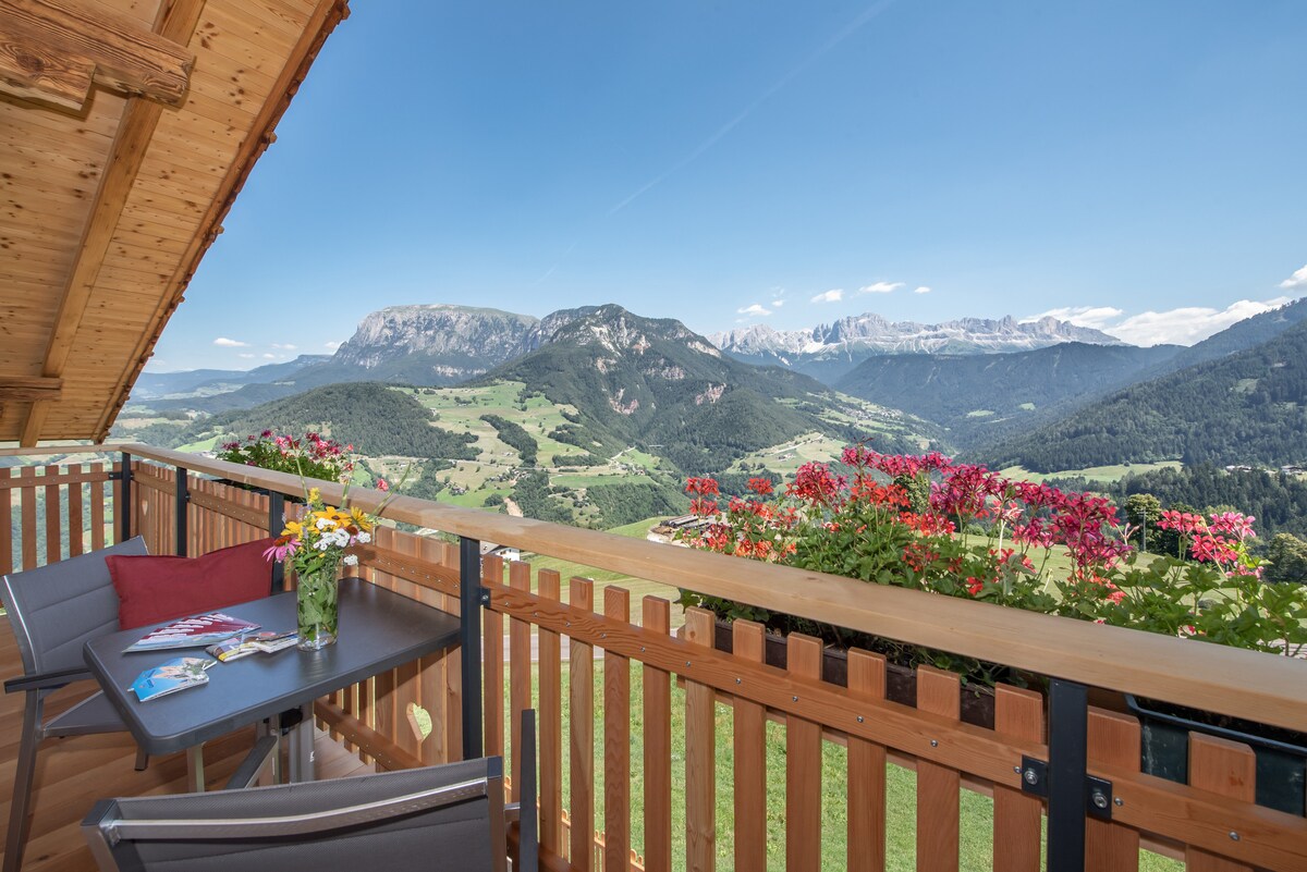 A private balcony is visible, featuring a small table and two chairs. Colorful flowers in pots add vibrancy, while a stunning view of the Alps stretches across the horizon, showcasing lush greenery and mountainous landscapes under a clear blue sky.
