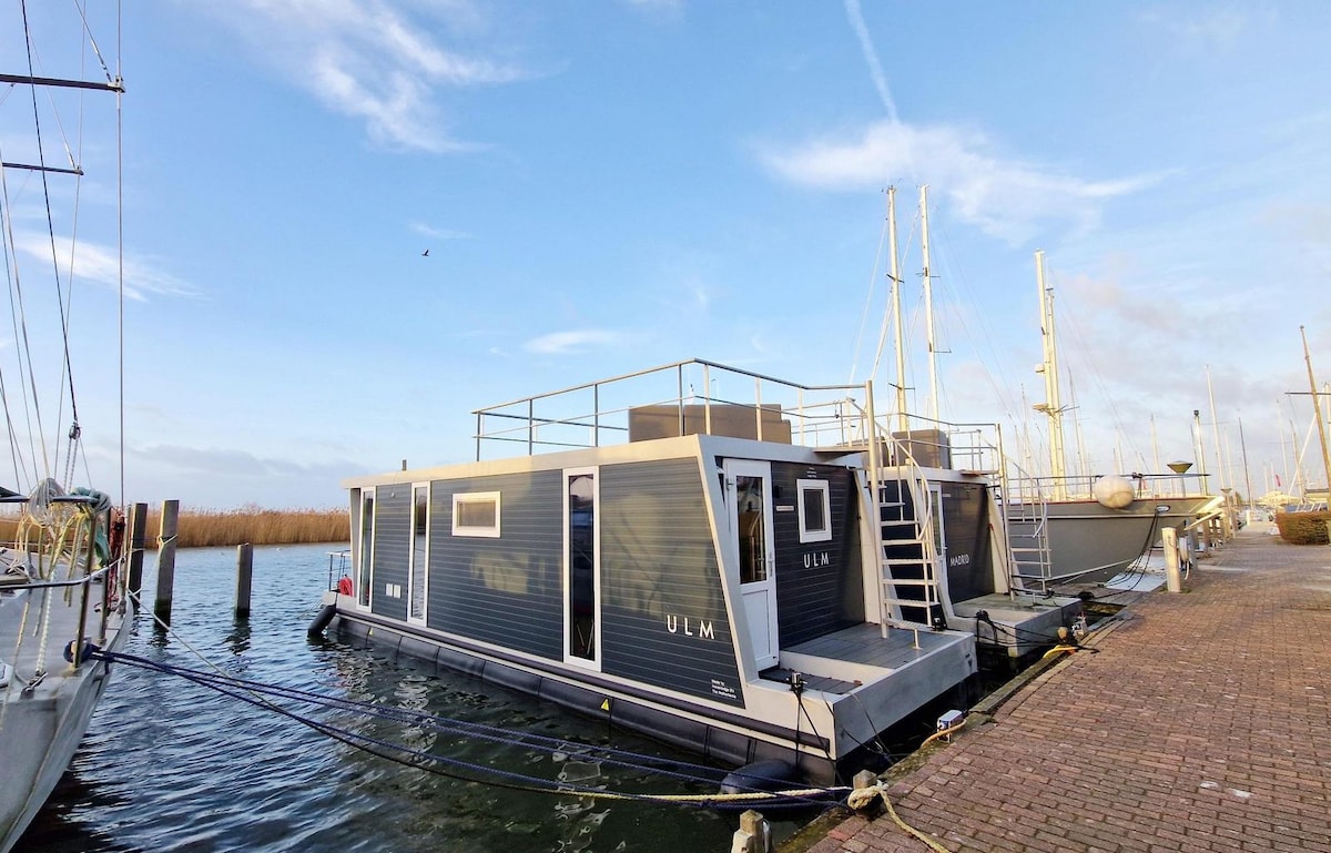 A modern floating lodge is moored at a dock, featuring a sleek design with large windows and a spacious rooftop terrace. The calm water surrounds the lodge, and other boats are visible in the background, enhancing the waterfront setting.