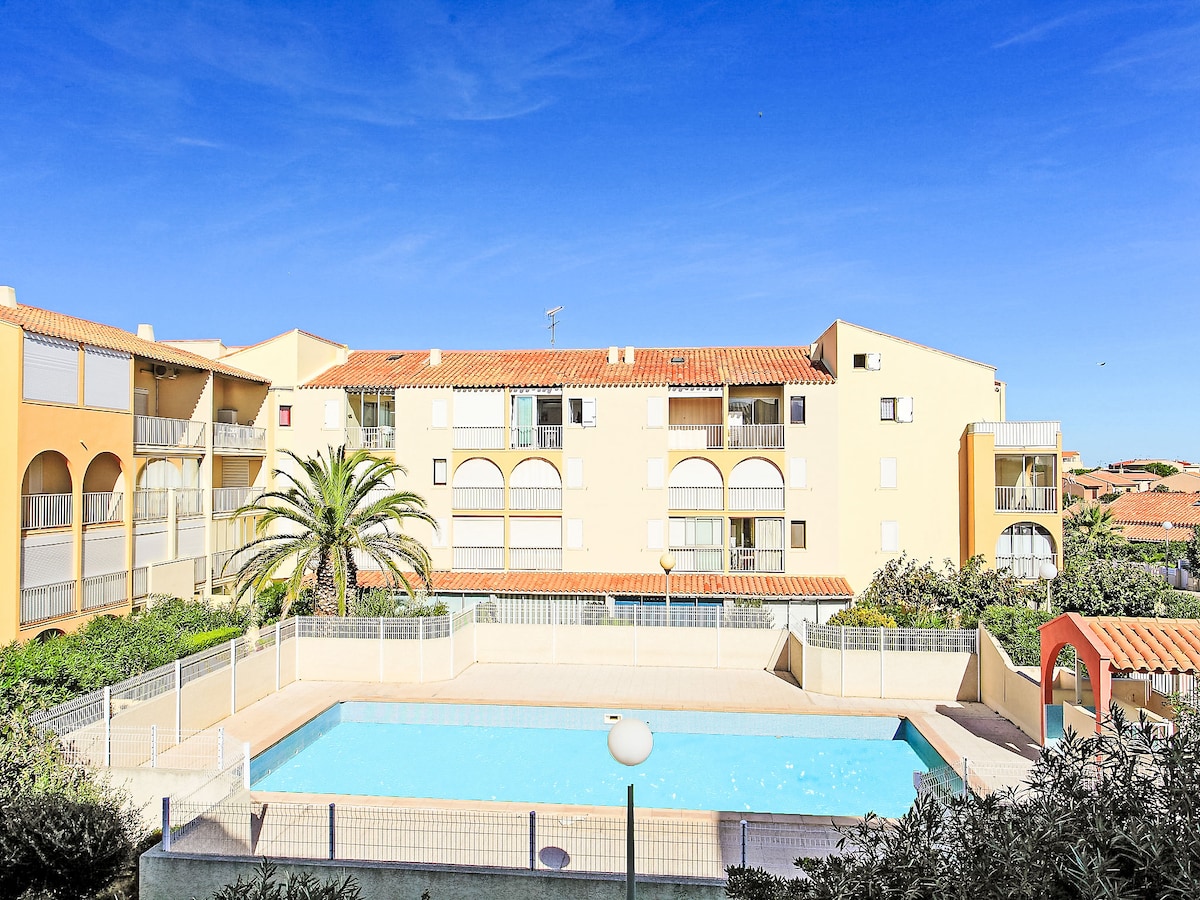 A view of the outdoor swimming pool is presented, framed by palm trees and surrounded by a low wall. The two-story building features a terracotta roof and large, arched windows. Clear blue skies complement the bright, inviting setting.