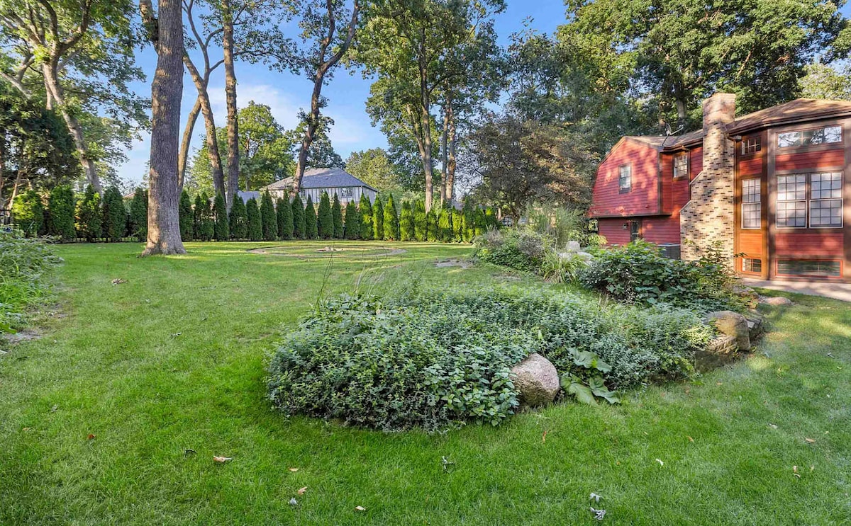 A spacious backyard is surrounded by tall trees and lush landscaping, featuring a well-maintained lawn. A quaint red building is visible, adding charm to the natural setting. The area invites relaxation and enjoyment of the outdoors.