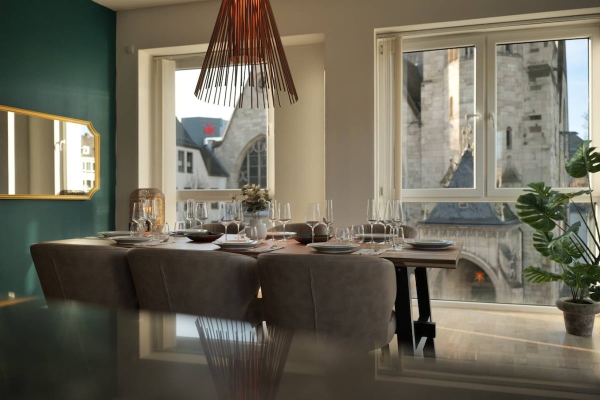 A dining area features a large wooden table set for eight, complemented by soft, cream-colored chairs. Sunlight streams through expansive windows, framing views of nearby historic architecture. A potted plant and decorative flower arrangement enhance the inviting ambiance.