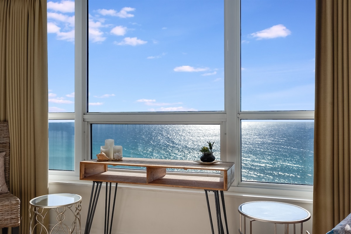 A bright living area features large windows offering expansive views of the Gulf of Mexico. Two round side tables are positioned beside a wooden console table, which is accented with decorative items. Soft natural light illuminates the space.