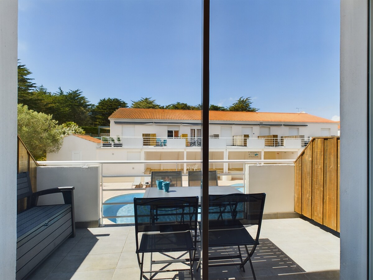 A balcony view reveals a communal heated swimming pool and a well-maintained courtyard, surrounded by buildings with terracotta roofs. Two black chairs and a small table are positioned for outdoor relaxation, with greenery providing a natural touch to the scene.