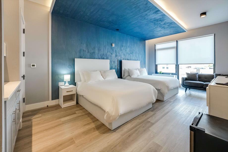 A spacious sleeping area is highlighted by two large beds with crisp white linens. A deep blue accent wall adds a modern touch, while natural light filters through the windows featuring roller shades. A cozy seating area and kitchenette complete the inviting atmosphere.