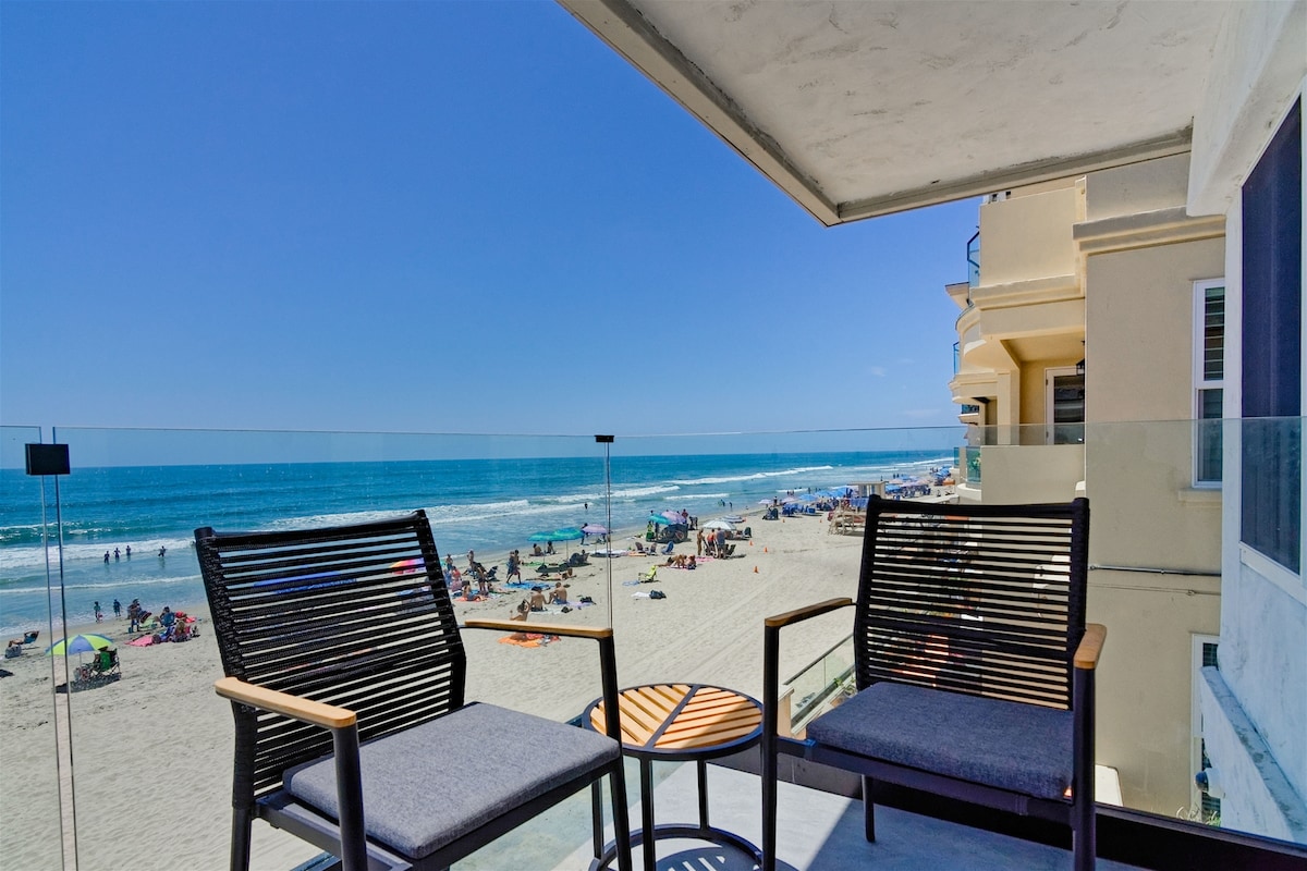 An oceanfront deck features two stylish chairs paired with a small table, offering unobstructed views of the sandy beach and surf. The scene includes sunbathers and umbrellas dotting the shore under a clear blue sky.