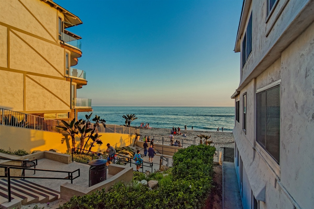 A view from the property shows a pathway leading to the beach, with steps and greenery lining the sides. The ocean is visible in the distance, complemented by a clear sky at sunset. People can be seen enjoying the shoreline.