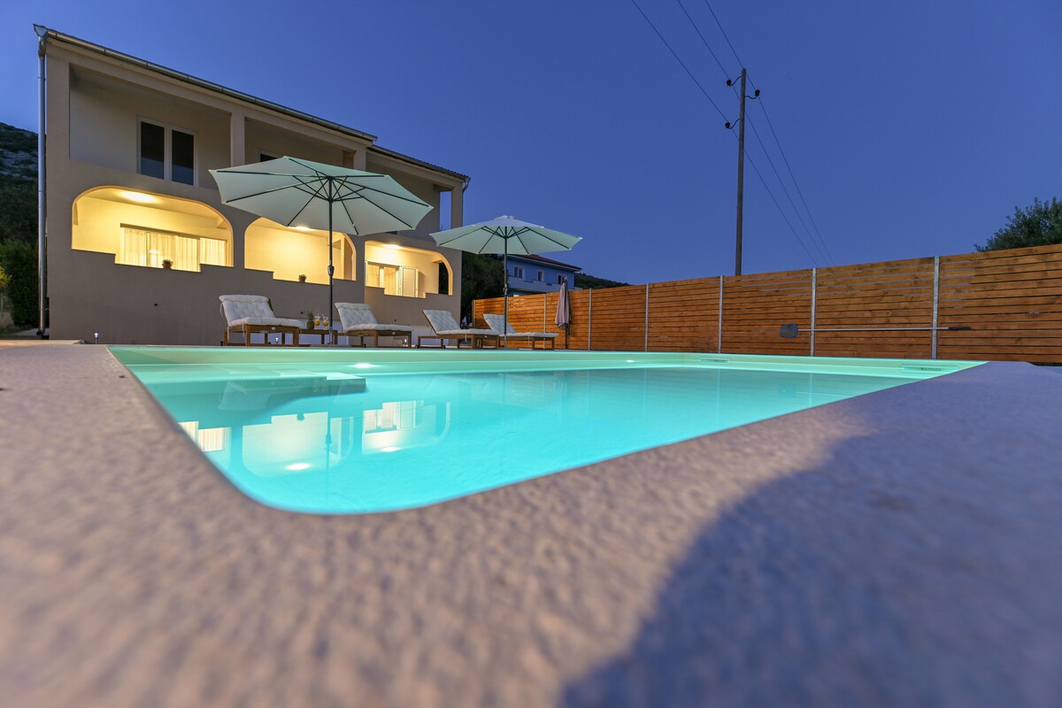 A private pool is highlighted with gentle lighting, reflecting a serene turquoise hue. Sun loungers are positioned nearby, accompanied by large umbrellas for shade. The backdrop features a two-story house with large windows, showcasing a welcoming atmosphere in the evening light.