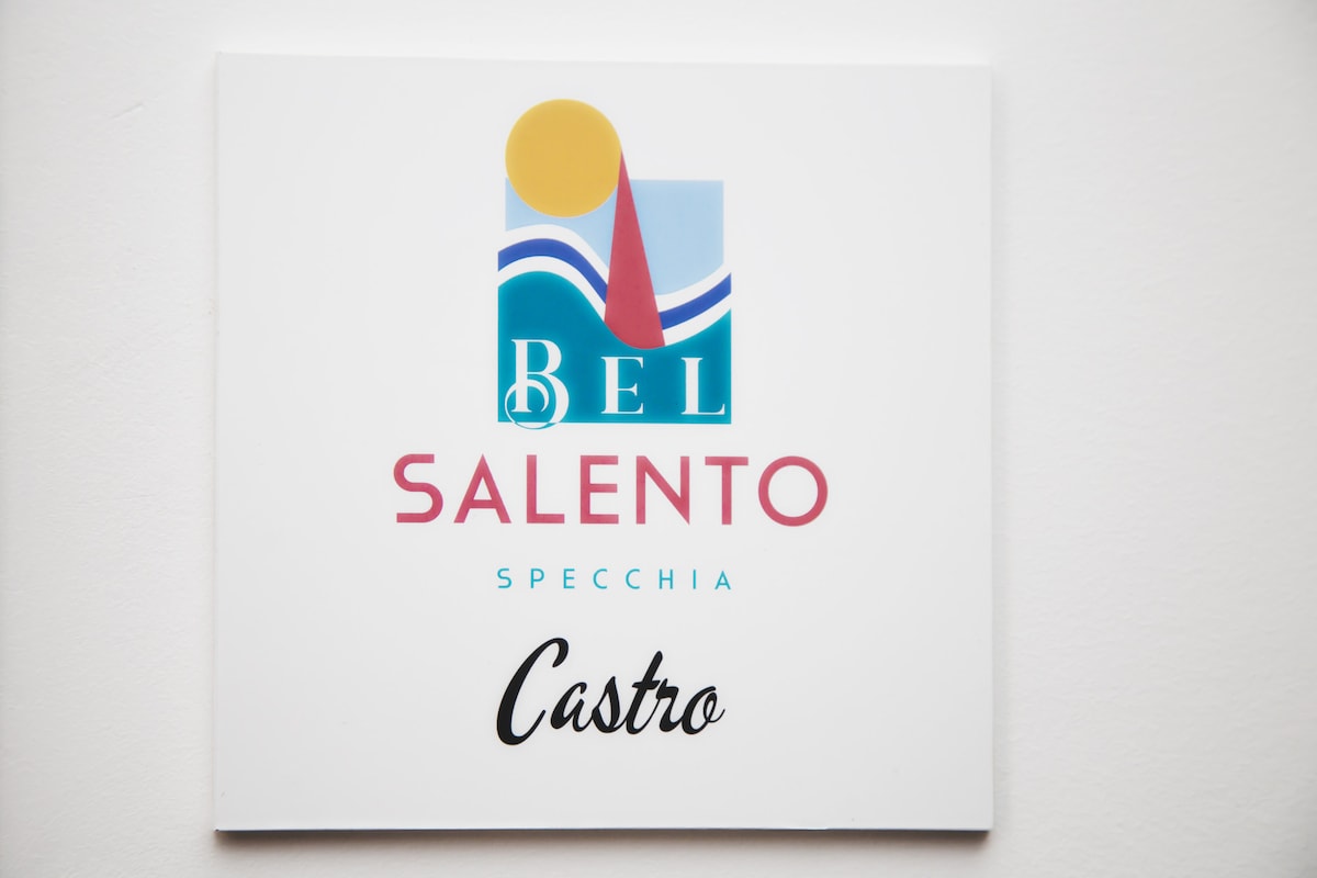 A colorful sign is displayed, featuring a logo for 'Bel Salento' with 'Castro' elegantly scripted below. The design includes a sun, waves, and a vibrant red triangle, creating a welcoming representation of the Salento region.