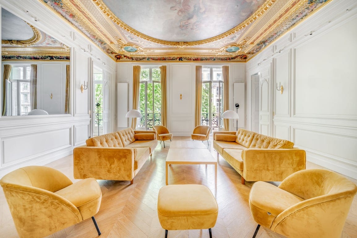 A spacious living area features a high, ornately painted ceiling from the 18th century. Soft, golden sofas and armchairs are arranged around a central table, with large windows allowing natural light to fill the room. A mirror enhances the sense of space.