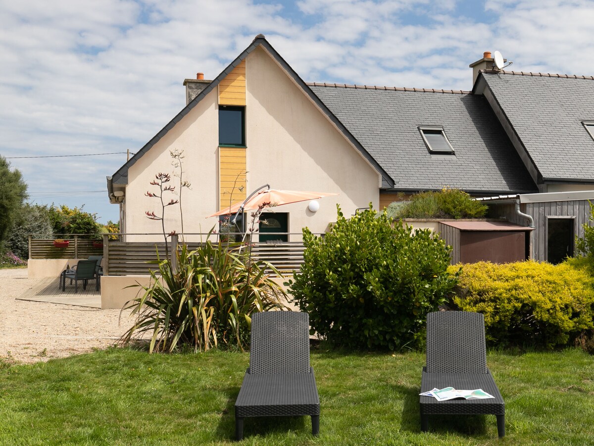 The exterior of the house features a modern design with a combination of natural elements and sleek lines. Two deck chairs are placed on the well-maintained lawn, surrounded by vibrant greenery and colorful plants. A terrace with seating is visible in the background.
