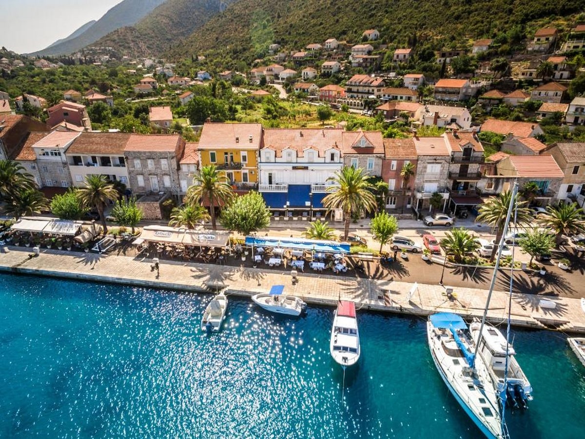 A waterfront view captures the charming streets of Trpanj, lined with trees and small-scale boats moored along the pier. Colorful buildings with terracotta roofs create a picturesque backdrop, while the surrounding hills provide a serene natural setting.
