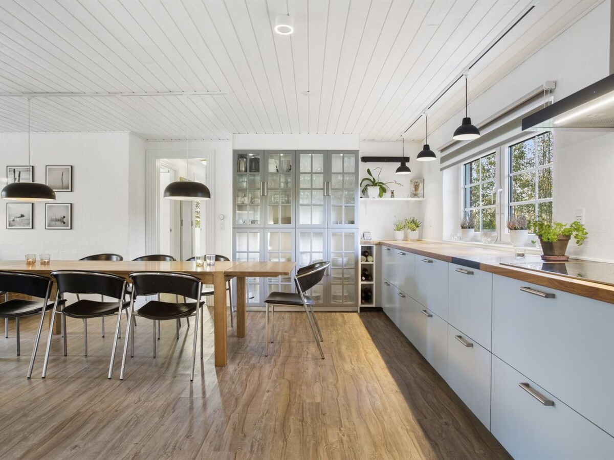 A modern kitchen features sleek cabinetry and a spacious wooden dining table surrounded by black chairs. Large windows provide natural light, illuminating the countertops and highlighting the organized shelves with glass doors.