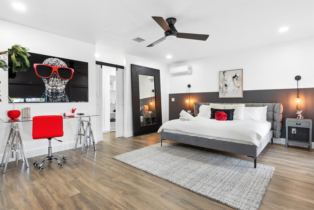 A spacious bedroom is featured, showcasing a king bed with white linens and decorative pillows. A large wall-mounted art piece of a dog in sunglasses is accompanied by a stylish desk with a bright red chair. Light hardwood flooring enhances the modern design.