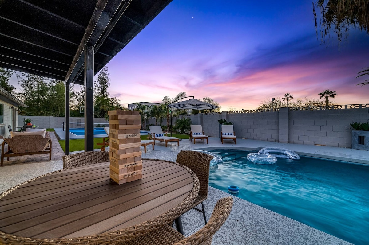The outdoor space features a dining table surrounded by woven chairs, positioned near a sparkling pool. A colorful sunset creates a serene backdrop, while loungers with cushions are placed beside the water. A game of Jenga is set on the table, adding to the inviting atmosphere.