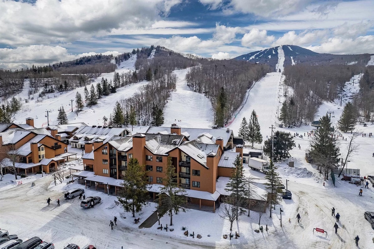 A panoramic view of Pico Mountain's ski area showcases snow-covered slopes and neatly aligned trees. The building, featuring a warm-toned facade, is situated in the foreground, emphasizing its proximity to the lifts. Skiers can be seen enjoying the slopes, illustrating the winter activity.