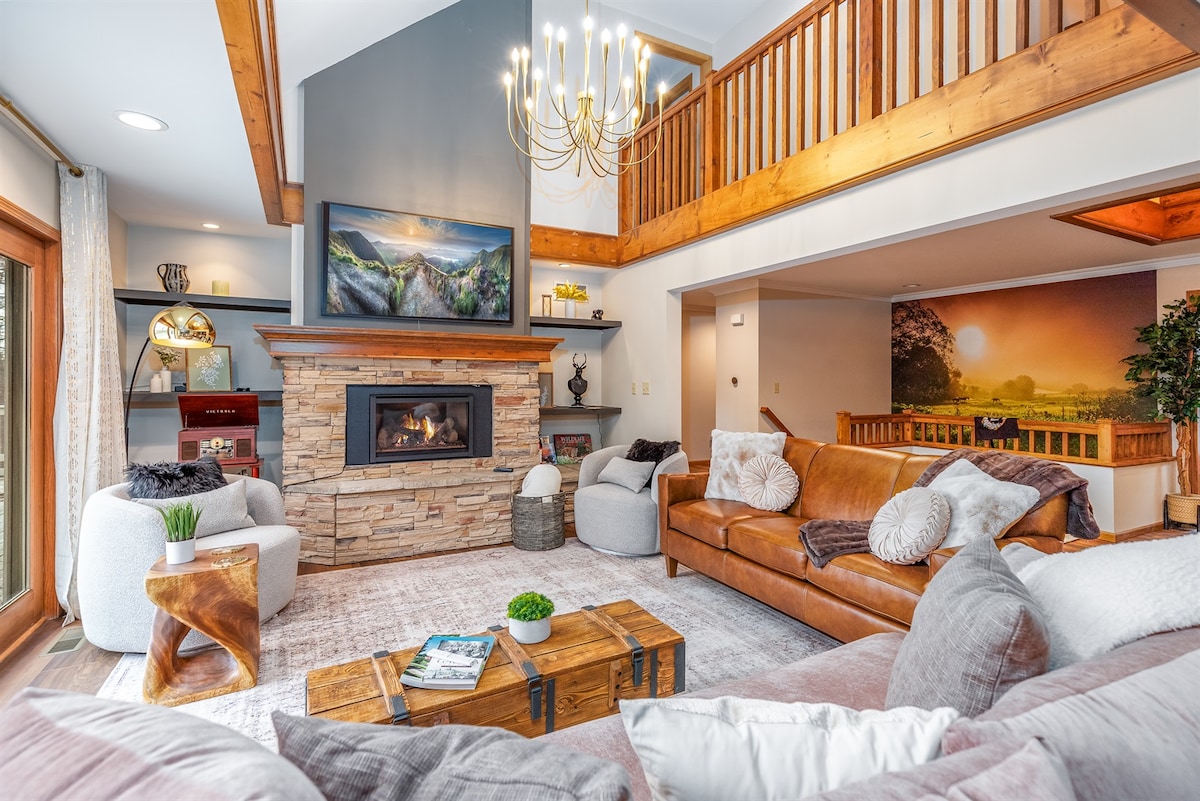 A spacious living area features a stone fireplace framed by a wooden mantel and a large artwork depicting mountains. Comfortable seating includes a leather sofa and plush armchairs, arranged around a central coffee table. Natural light enters through sliding glass doors, enhancing the inviting atmosphere.