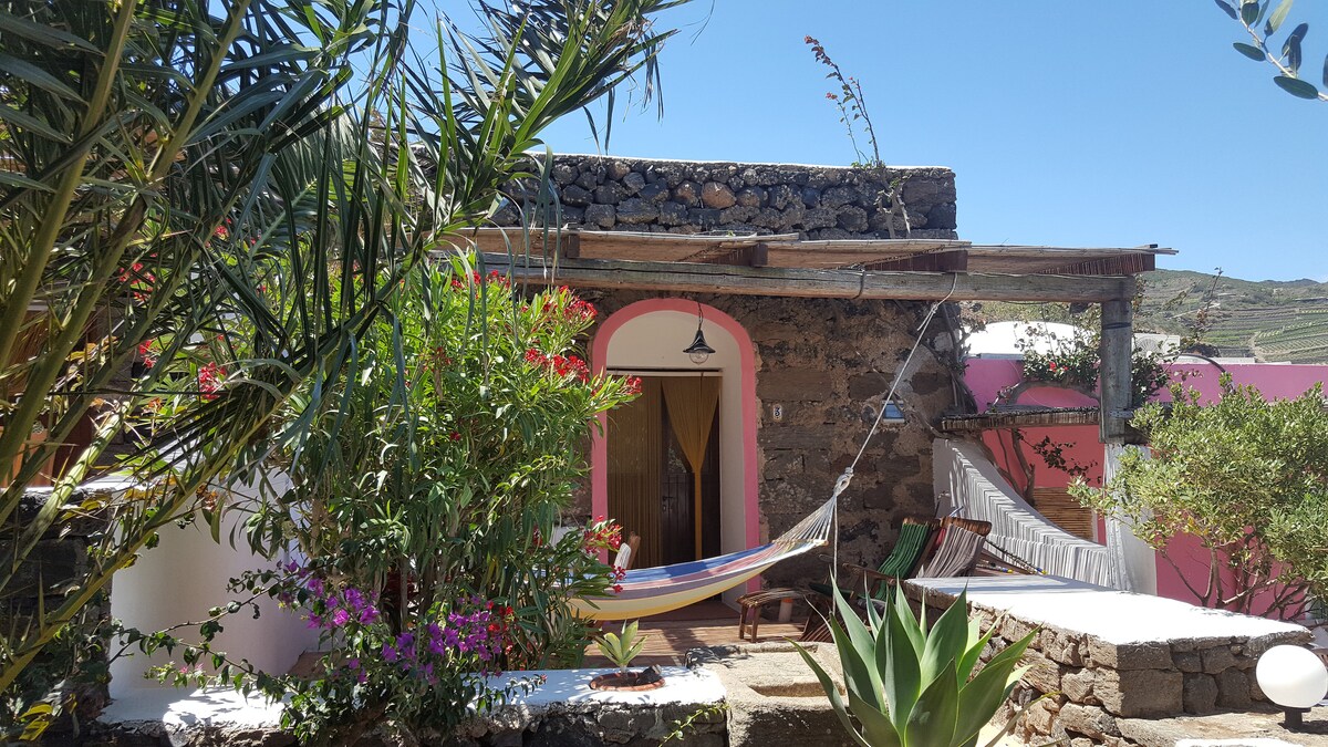 A stone structure with a warm entrance is framed by lush greenery and colorful flowers. A hammock is suspended between supportive beams, inviting relaxation. The setting features a blend of natural elements with bright walls, showcasing the tranquil atmosphere of the surrounding countryside.
