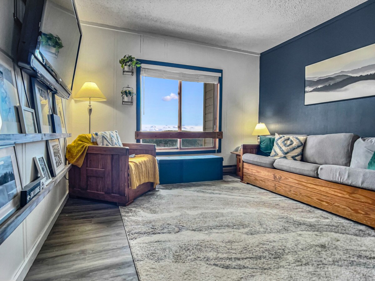 A cozy living room features a large window showcasing mountain views. A stylish sectional sofa and a wooden bench provide comfortable seating. A flat-screen TV is mounted on the wall, and a soft area rug complements the space, adding warmth and inviting ambiance.