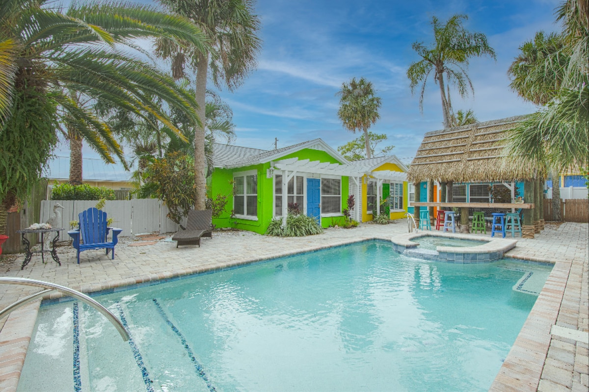 A vibrant pool area is surrounded by tropical palm trees and colorful cottages. The clear, inviting pool is accompanied by a hot tub and a thatched-roof tiki bar, providing a relaxed atmosphere. Paved stones create an open layout, enhancing the inviting outdoor space.