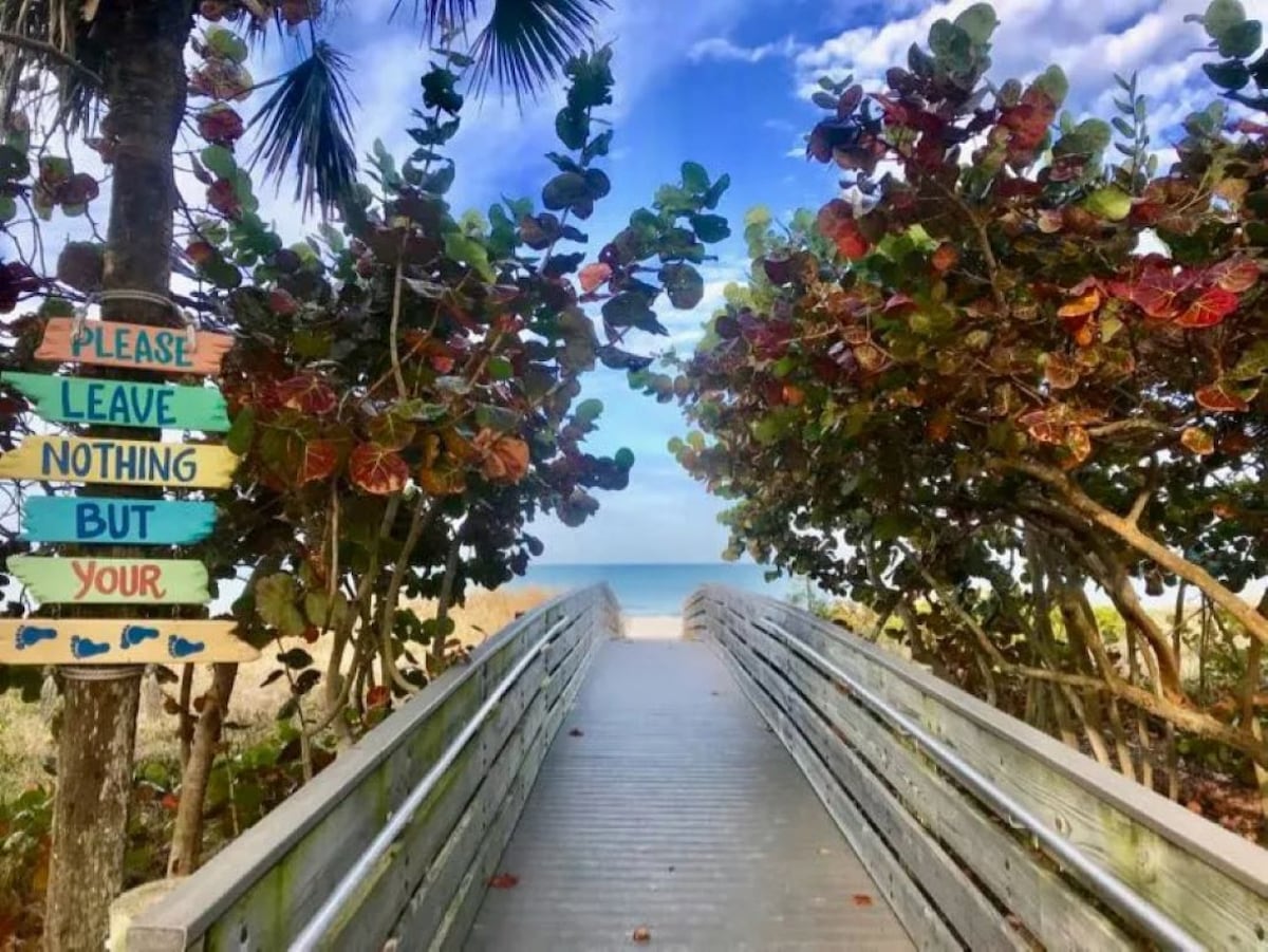 A wooden boardwalk is framed by lush greenery and colorful foliage leading towards a serene beach. A sign reading 'Please Leave Nothing But Your' is visible on the left. The tranquil ocean is visible in the distance under a partly cloudy sky.