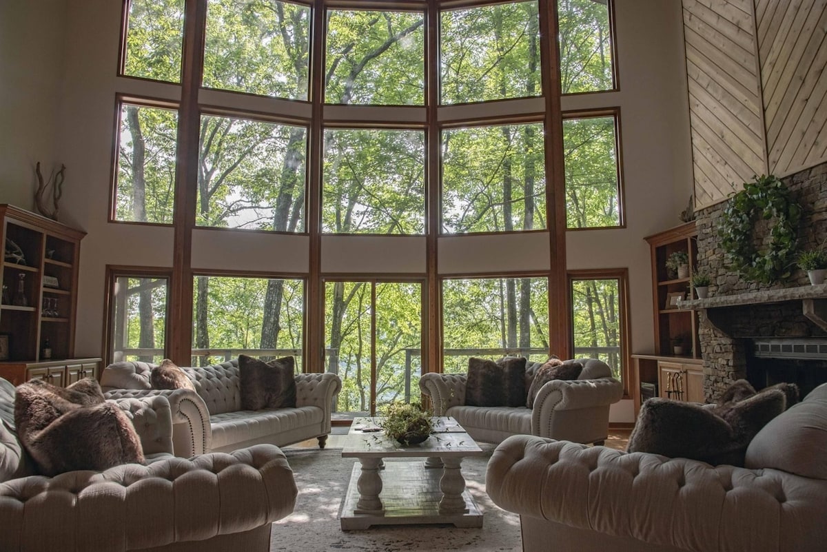 A spacious living area features a two-story wall of windows that allows natural light to fill the room. Comfortable seating is arranged around a central coffee table, complemented by soft throw pillows. Lush greenery from the surrounding trees is visible through the windows.